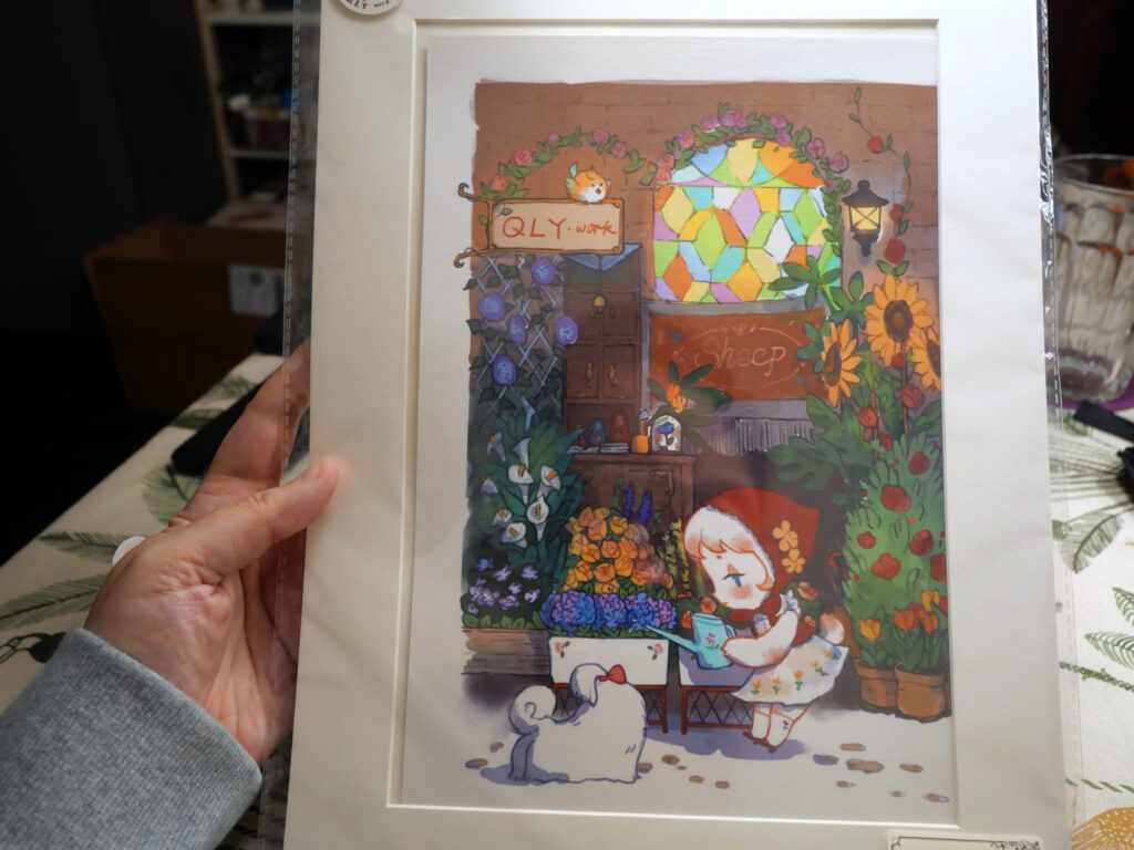 A colourful print shown of a anthropomorphic sheep wearing a white dress with yellow and green flower trim and a red headdress with 2 yellow flowers attached to it. She is holding a blue watering can and is watering some pretty purple hydrangea while a little white dog with a pink bow is look at her activities. 

She is running a flowershop with many pretty flowers like sun flowers, roses, tulips and calla lilies.