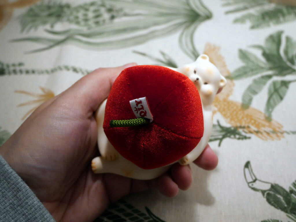 A pin cushion shaped in the form of a white bear while on his tummy a red giant stuffed apple is placed.