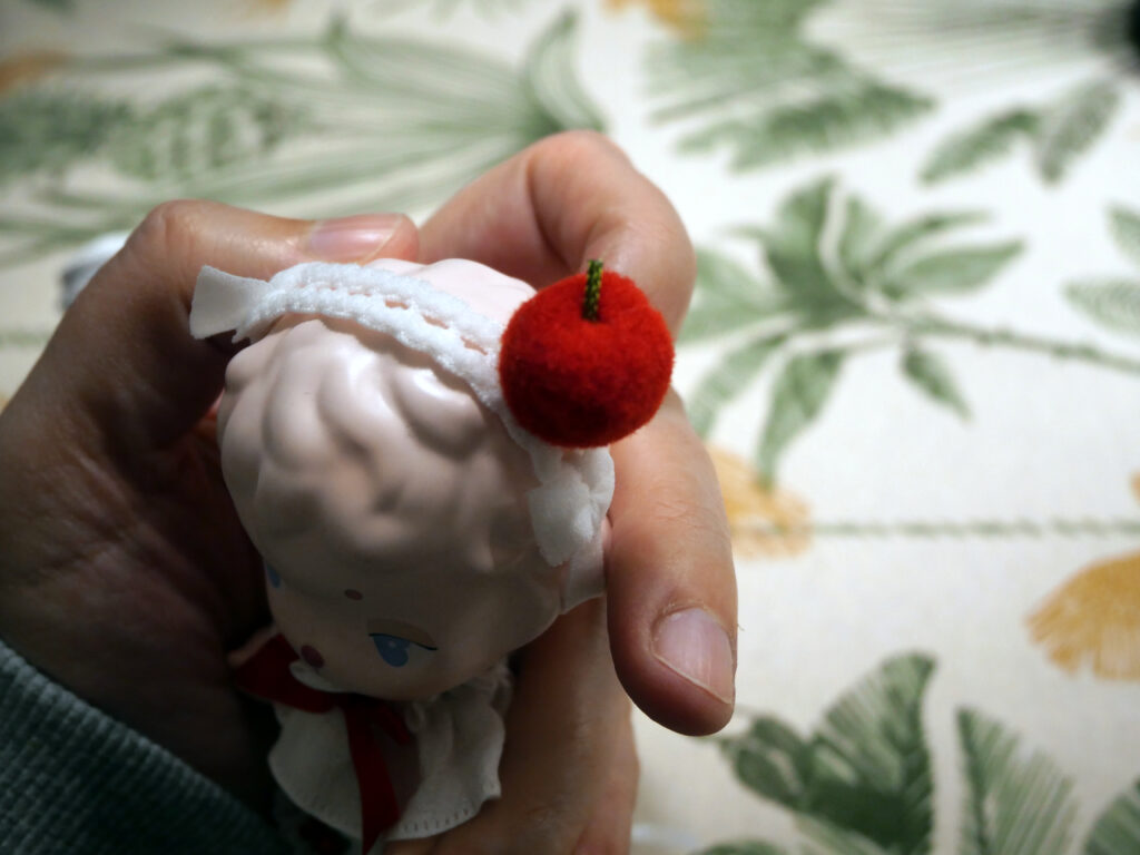 Close up of the red apple with stem attached to the frilly white headdress worn by a pink sheep doll