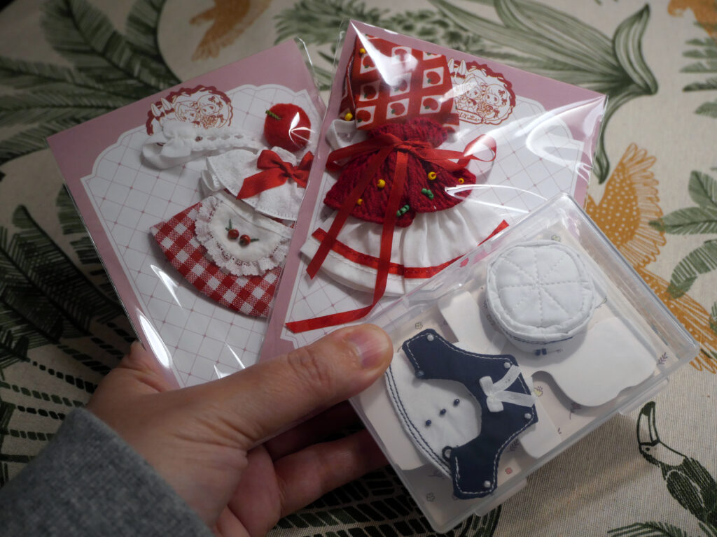 Three outfits still in their original packaging. On the left a red checkered dress, with a white frilly apron decorated with two apples, a frilly collar and a frilly headdres with an apple. 

In the middle is a red riding hood outfit, with red trimmings, a red caplet and a red headscarf with images of apples printed on them.

On the right is a white sailor outfit with blue thread trimmings, a blue collar with a white bow and white pillbox hat. 